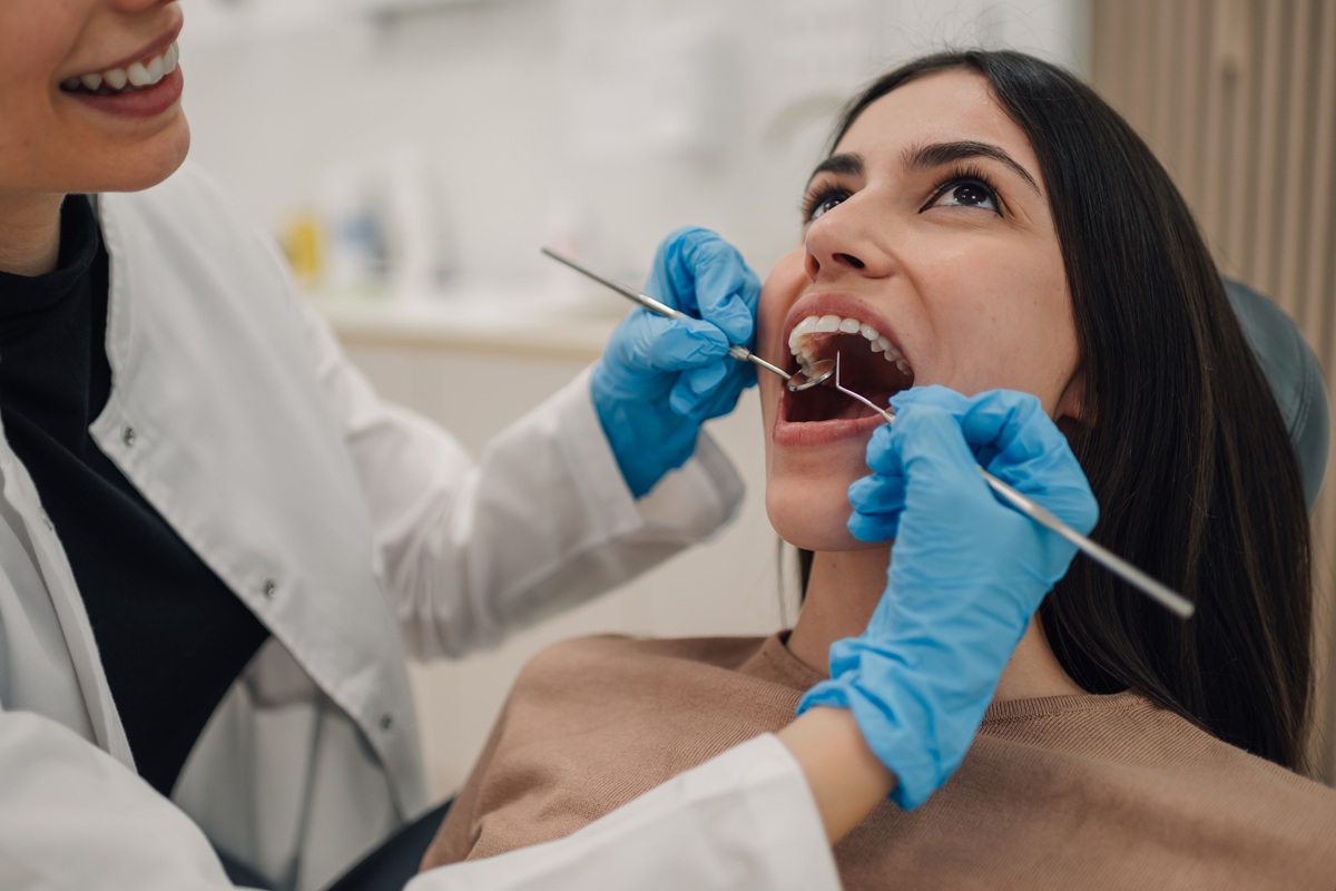 Female dentist wearing blue gloves examining teeth of female patient with dental tools in dental clinic, performing oral hygiene and teeth checkup