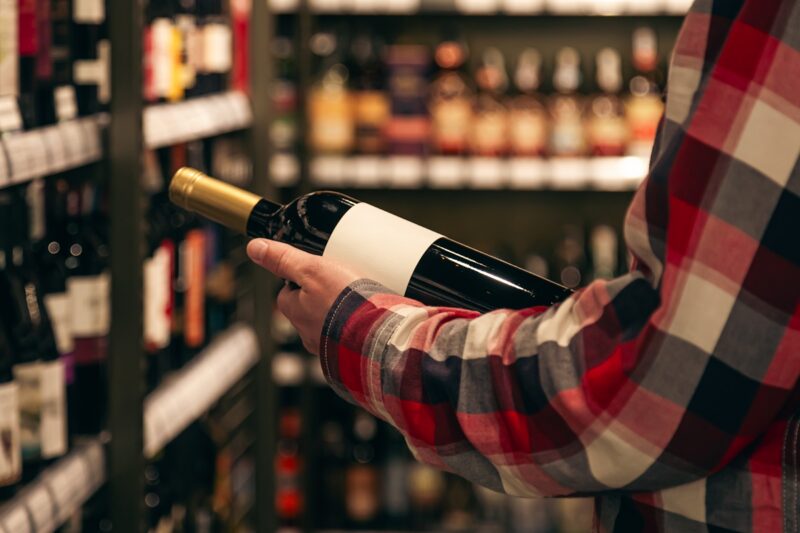 A man holding a wine bottle in a liquor wine shop, choosing the right wine from all the variations of wine bottles on the shelves.
