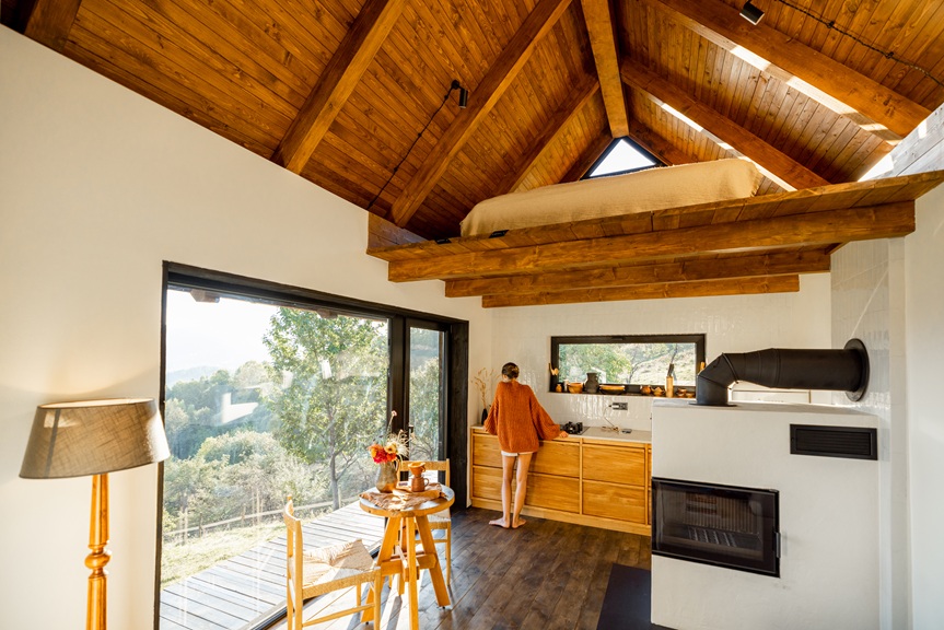 A woman in an orange sweater stands in a rustic kitchen with wooden cabinets, enjoying the tranquil morning light. The warm interior features a fireplace, large windows, and a relaxed atmosphere