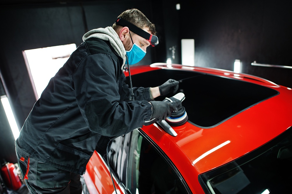 Car detailing concept. Man in face mask with orbital polisher in repair shop polishing roof of orange suv car.